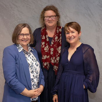 Three women smiling and standing together indoors, dressed in professional attire with a plain textured wall background.